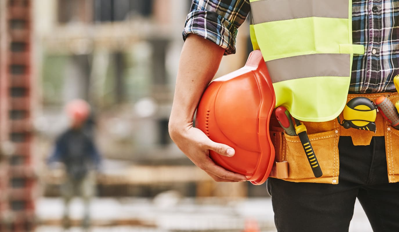 Imagen de un obrero de la construcción que sostiene un casco mientras lleva también un chaleco de obra y un cinturón de herramientas.